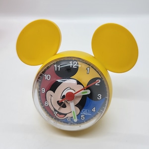 May include: Yellow alarm clock with Mickey Mouse's face on the clock face. The clock has Mickey Mouse ears on the top. The clock face is multi-colored with red, yellow, blue, and black. The clock face has black numbers and the text "G.I.A. International" at the bottom.