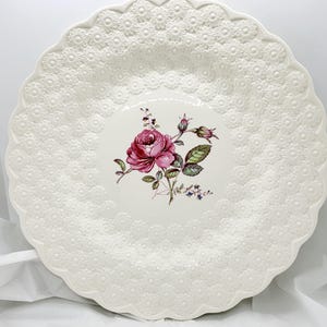 May include: A white ceramic serving plate with a scalloped edge and embossed floral pattern. The center features a pink rose illustration with green leaves and buds. A decorative piece for serving food.