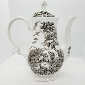 Peut inclure: Cafetière en céramique blanche avec couvercle, ornée d'une scène pastorale en noir et blanc. Le motif comprend un chasseur, une maison, des arbres et des oiseaux. La poignée et le bec sont incurvés.