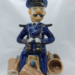 May include: A blue and white ceramic figurine of a sailor wearing a captain's hat and holding binoculars. The sailor is sitting on a barrel with four small ceramic mugs attached to the sides.