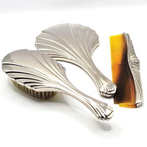 May include: A silver-toned vanity set consisting of a hairbrush with light-coloured bristles, a hand mirror, and a comb with a tortoiseshell-patterned finish. Each piece features a matching fan-shaped, ribbed design on the metallic handles and backs.