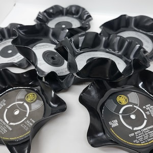 May include: Six black vinyl record coasters with a ruffled edge. The coasters feature the labels of various albums, including "Philadelphia Freedom" by Elton John and "Step Into Christmas" by Elton John.