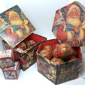 May include: A collection of decorative boxes with festive Christmas imagery. The boxes display illustrations of Santa Claus, children, and other seasonal motifs. One box holds Christmas baubles. The boxes are red and gold.
