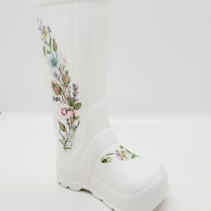 May include: White ceramic boot vase with a floral design. The boot is decorated with a variety of flowers and leaves in shades of pink, blue, green, and yellow.