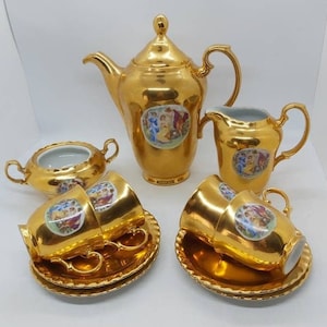 May include: A gold-colored tea set with a teapot, creamer, sugar bowl, and cups with saucers. Each piece has a decorative oval image with figures. The set has a shiny, reflective surface and a classic design.