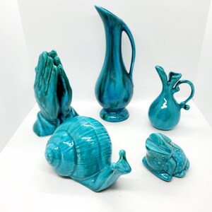 May include: Five teal blue ceramic figurines: a pair of hands clasped in prayer, a snail, a frog, and two pitchers. The pitchers have different shapes and designs.