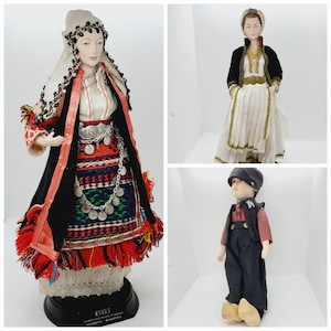 May include: Three porcelain dolls dressed in traditional folk costumes. The dolls are wearing colorful clothing with intricate embroidery and accessories. The dolls are standing on a white background.