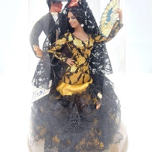 May include: A decorative figurine set inside a clear plastic dome. The set features two figures: a woman in a gold and black lace dress with a fan, and a man in a black suit and hat. The woman is wearing a black lace veil.
