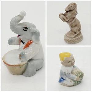 May include: Three porcelain figurines: a gray elephant playing a drum, a brown horse standing on its hind legs, and a yellow-haired gnome holding a green and brown object.