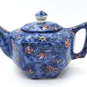 May include: Blue ceramic teapot with a floral pattern. The teapot features a lid with a small knob and a curved handle. The floral design includes pink, yellow, and white flowers and green leaves. The teapot is a decorative item for serving tea.