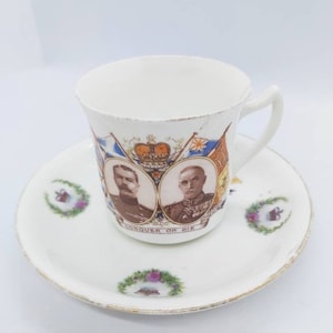 May include: A white porcelain teacup and saucer with a gold rim. The cup features a portrait of two men in military uniform, with the text "Conquer or Die" below. The saucer has a floral design with a crown.