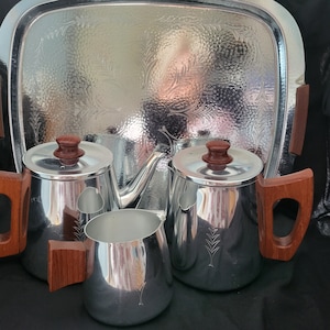 May include: A vintage silver tea set with a matching tray. The set includes a teapot, creamer, and sugar bowl, all with wooden handles and lids. The tray has a decorative embossed floral pattern.