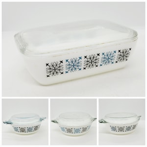 May include: A white rectangular Pyrex baking dish with a clear glass lid. The dish has a decorative band of black and blue snowflake-like designs. The lid is slightly curved and the dish has a gold trim.