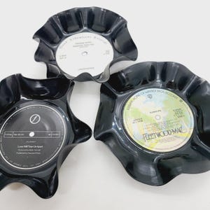 May include: Three black, wavy-edged bowls made from recycled vinyl records. Each bowl features a different record label, including "Teenage Kicks" by Undertones and "Rumours" by Fleetwood Mac. A unique, eco-friendly home decor item.
