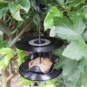 May include: A black vinyl record bird feeder with a black metal hanger and black ribbon. The feeder is filled with birdseed and is hanging from a tree branch.