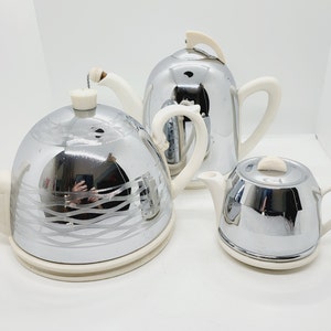 May include: A set of three silver teapots with white lids and handles. The teapot on the left has a decorative design on the side. The teapot in the middle has a tall, slender spout. The teapot on the right has a rounder shape.
