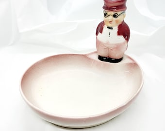 以下が含まれることがあります： ヴィンテージの陶器の皿で、フィギュアのトッパーが付いています。 皿はハート型で、ピンク色の縁と白い内側があります。 トッパーは、バーガンディのスーツとメガネをかけた男性の小さな置物です。