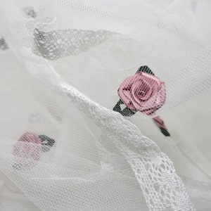 May include: White tulle fabric with pink fabric roses. The tulle has a delicate lace trim.