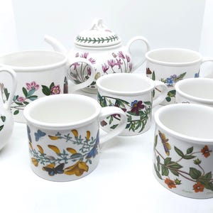 May include: A white ceramic tea set comprising a teapot, a milk jug, and several mugs. The set is adorned with colourful floral and butterfly patterns. The teapot has a lid with a floral design. The mugs have handles and various floral designs.