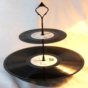 May include: A two-tiered serving stand made from vintage vinyl records. The black records are held together by a black metal stand with a decorative handle. The labels on the records are visible.