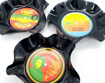 Może przedstawiać: Trzy czarne podkładki pod kubki z winylu z Bob Marleyem i The Wailers. Podkładki mają kształt kwiatu z falowanym brzegiem. Środek każdej podkładki przedstawia inną okładkę albumu w czerwono-żółto-zielonej kolorystyce.