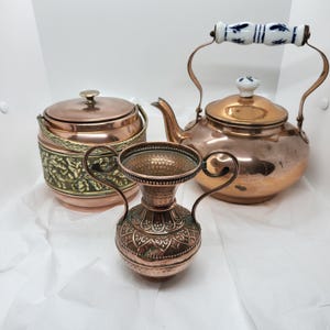 Peut inclure: Trois articles de cuisine en cuivre : un petit vase avec des poignées, un récipient avec un couvercle et un motif floral en laiton, et une théière en cuivre avec une poignée blanche et bleue. Les articles sont disposés sur une surface blanche.