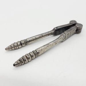 May include: A silver metal nutcracker with a textured design and a hinged jaw. The nutcracker has a long, thin handle with a spiral design.