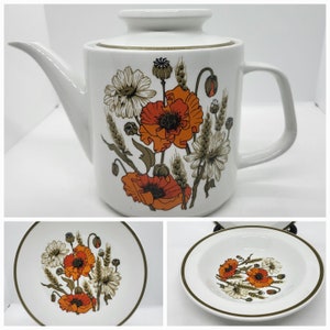 May include: A white teapot with a floral design featuring orange poppies and white daisies. The teapot has a lid and a handle. The design is on a white background with a gold trim around the top.