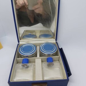 May include: A vintage vanity set in a blue case, featuring a mirror, two jars with blue lids, and two clear glass bottles with blue stoppers. The set has a silver clasp and a white interior.
