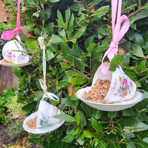 May include: Three teacups with saucers are hung from a tree branch with pink ribbon. The teacups are filled with birdseed and have the word "Mother" written on them.