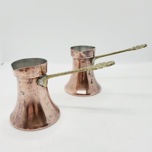 May include: Two copper Turkish coffee pots with brass handles. The pots have a hammered texture and are a traditional design.