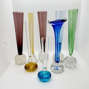 May include: A set of eight glass bud vases with round bases. The vases are in various colours, including purple, yellow, clear, blue, and green. The bases of the vases are textured with small bubbles.