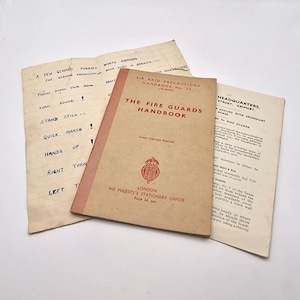 May include: An open vintage handbook titled "The Fire Guards Handbook" with red and white text, and a handwritten sheet with German phrases. The handbook is from His Majesty's Stationery Office, London.
