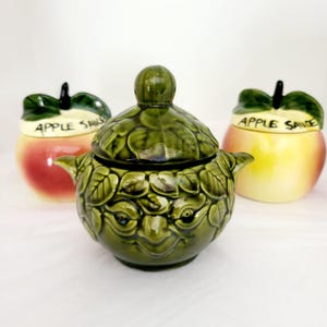 Könnte beinhalten: Drei Keramik-Apfelsaucenbehälter. Zwei sind apfelförmig mit der Aufschrift "Apple Sauce". Einer ist grün, hat einen Deckel und ist wie ein Gesicht mit Blättern geformt.