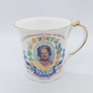 May include: White commemorative mug with gold trim, featuring a portrait of Queen Elizabeth II. The mug is decorated with floral and ribbon motifs, and text that reads "To Commemorate the 90th Birthday".