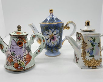 May include: Three decorative teapots with floral designs. The teapot on the left is green with orange and pink flowers. The teapot in the center is blue with white flowers. The teapot on the right is white with gold accents and blue flowers.