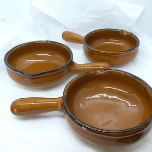 May include: Three brown ceramic soup bowls with handles. The bowls have a glossy finish and a dark brown rim. The handles are curved and attached to the side of each bowl. The bowls are arranged on a white surface.
