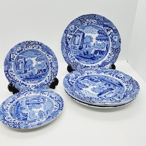 May include: A collection of blue and white ceramic plates, each displaying a landscape scene. The plates showcase a detailed illustration of a classical setting with trees, buildings, and figures. The plates are on black stands.