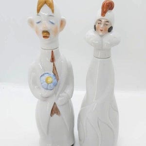 May include: Two white ceramic bottles with painted faces. One has a man's face with closed eyes, a brown mustache, and a blue flower. The other has a woman's face with a hat and a ruffled collar. Decorative bottles.