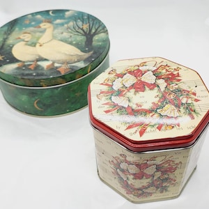 May include: Two round vintage tin boxes with festive designs. The larger box has a green background with a white bird and stars. The smaller box has a red and white background with a wreath of red and white bells and bows. The text on the smaller box reads "so mit  schil les Haar".