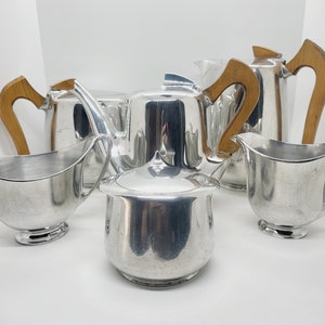 Peut inclure: Un ensemble de cinq pièces de service à thé en argent, comprenant une théière, un pot à crème, un sucrier et deux tasses. La théière et le pot à crème ont des poignées en bois.