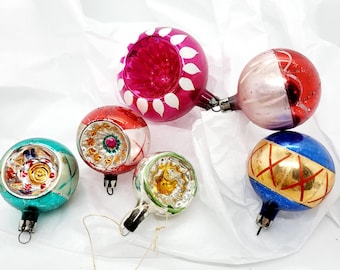 May include: Six vintage glass Christmas ornaments with various designs. The ornaments are in shades of pink, red, blue, green, and white. Some ornaments have geometric patterns, while others have floral designs.