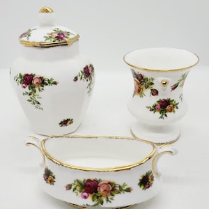 Peut inclure: Ensemble de trois pièces en porcelaine blanche ornées d'un motif de roses. L'ensemble comprend un pot avec couvercle, un petit vase et un plat à anses, tous bordés d'or. Le motif de roses présente des fleurs bordeaux, roses et jaunes avec des feuilles vertes.