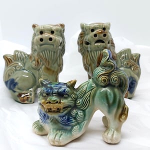 May include: Three ceramic Shisa statues in shades of green, blue, and brown. The statues have detailed features, including expressive faces and ornate tails. The Shisa are arranged on a white surface, showcasing their traditional Okinawan design.
