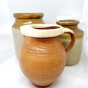 May include: Three ceramic pottery pieces, a brown jug with a white rim and handle, a brown and beige jar, and a brown and beige jar with a lid.