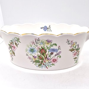 Könnte beinhalten: Eine ovale Servierschale in Weiß mit gewelltem Rand und goldenem Rand. Sie hat florale Designs in Rosa, Blau, Gelb und Grün. Zwei kleine Griffe befinden sich auf beiden Seiten der Schale.