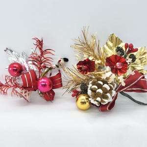 May include: Two decorative holiday floral arrangements featuring red and pink ornaments, silver and gold metallic leaves, pine needles, and red ribbons. The left piece includes a small snowman figure, while the right piece features a pinecone.