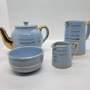 Könnte beinhalten: Ein blaues und goldenes Teeservice mit Teekanne, Milchkännchen, Zuckerdose und einer passenden Tasse. Das Set hat ein zartes goldfarbenes Streifenmuster.