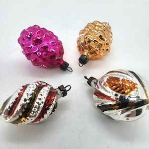 May include: Four vintage Christmas ornaments in various shapes and colors. One is a bright pink, textured shape. Another is gold, and two are silver with red and gold accents. Each has a black metal cap and a loop for hanging.
