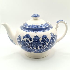 Könnte beinhalten: Weiße Keramik-Teekanne mit blauem Willow-Muster. Die Teekanne hat einen abgerundeten Körper, einen gebogenen Ausguss und einen Henkel. Der Deckel ist mit einem kleinen Knopf versehen. Das blaue Muster zeigt eine Landschaft mit Gebäuden und Bäumen.